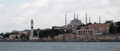 The image displays a coastal cityscape featuring a prominent mosque with multiple minarets, set against a cloudy sky. In the foreground, there is a shoreline with ancient stone walls, a lighthouse, and several historic buildings.
