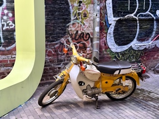 A vintage yellow and white Honda C70 motorcycle is parked on a brick pathway in front of a graffiti-covered brick wall. The background features colorful graffiti art in shades of red, green, and black. The motorcycle is positioned near a large, light green curved structure, possibly part of a sculpture or art piece. A metal pole with various stickers is also visible behind the motorcycle.
