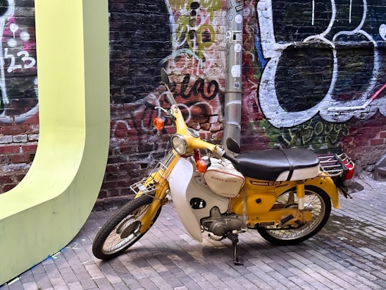 A vintage yellow and white Honda C70 motorcycle is parked on a brick pathway in front of a graffiti-covered brick wall. The background features colorful graffiti art in shades of red, green, and black. The motorcycle is positioned near a large, light green curved structure, possibly part of a sculpture or art piece. A metal pole with various stickers is also visible behind the motorcycle.