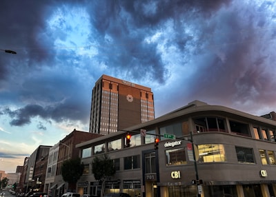 A cityscape featuring a tall building with the word 'TRUIST' on it, set against a dramatic sky filled with dark, swirling clouds. The street below shows various commercial buildings with signs like 'CBI' and 'delegator'. Traffic lights and street signs are visible in the foreground.