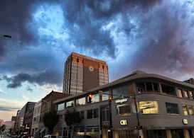 A cityscape featuring a tall building with the word 'TRUIST' on it, set against a dramatic sky filled with dark, swirling clouds. The street below shows various commercial buildings with signs like 'CBI' and 'delegator'. Traffic lights and street signs are visible in the foreground.
