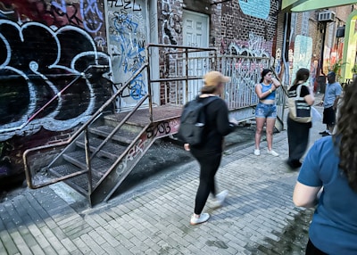 A group of people are gathered in an urban alley filled with colorful graffiti on the walls. Metal stairs lead up to a door, surrounded by vibrant street art and brick walls. Several individuals are engaged in different activities, with some walking past and others standing or talking.