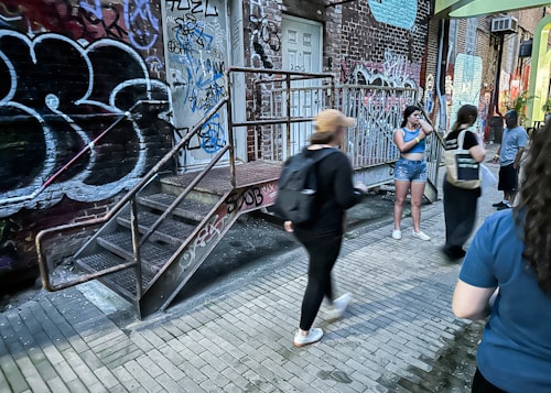 A group of people are gathered in an urban alley filled with colorful graffiti on the walls. Metal stairs lead up to a door, surrounded by vibrant street art and brick walls. Several individuals are engaged in different activities, with some walking past and others standing or talking.