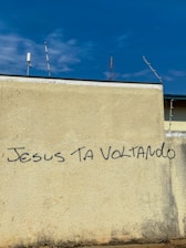 A beige wall with graffiti text reading 'JESUS TA VOLTANDO' in black letters. The top of the wall features a few antennas against a blue sky with scattered clouds.
