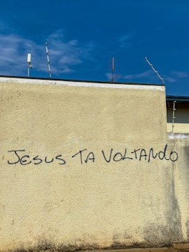 A beige wall with graffiti text reading 'JESUS TA VOLTANDO' in black letters. The top of the wall features a few antennas against a blue sky with scattered clouds.