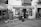 A charming storefront in black and white with a sign reading 'Casa Marieta' indicating a store selling artisanal beer, vermouth, typical sweets, and liqueurs. Two people are standing outside, seemingly looking at the display of bottles. There's a handwritten menu board, a small plant, and rustic barrels in front, all set on a cobblestone path.