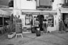 A charming storefront in black and white with a sign reading 'Casa Marieta' indicating a store selling artisanal beer, vermouth, typical sweets, and liqueurs. Two people are standing outside, seemingly looking at the display of bottles. There's a handwritten menu board, a small plant, and rustic barrels in front, all set on a cobblestone path.