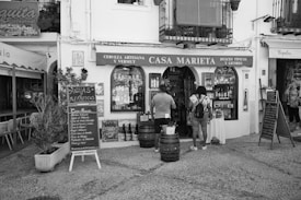 A charming storefront in black and white with a sign reading 'Casa Marieta' indicating a store selling artisanal beer, vermouth, typical sweets, and liqueurs. Two people are standing outside, seemingly looking at the display of bottles. There's a handwritten menu board, a small plant, and rustic barrels in front, all set on a cobblestone path.