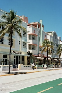A row of pastel-colored Art Deco buildings with geometric designs and large windows line the street. Tall palm trees sway gently in front of the buildings along a largely empty sidewalk under a bright blue sky.
