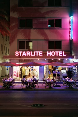 A neon-lit hotel facade with the name 'Starlite Hotel' prominently displayed in bright pink lights. The scene includes outdoor seating under patio umbrellas, where several people are dining. The building features a retro design with large windows, and the sidewalk dining area adds a lively atmosphere. There are palm tree shadows cast on the building, enhancing the tropical vibe.