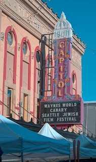 A historic theater building with an ornate facade features a large vertical sign displaying 'CAPITOL' in colorful letters. Below, a marquee lists events, including 'WAYNES WORLD,' 'CANARY,' and the 'SEATTLE JEWISH FILM FESTIVAL.' Several white tents are visible in the foreground, suggesting an outdoor event or market.