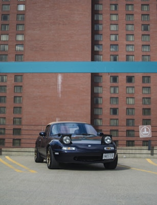 A black car with a license plate that reads 'RWDSTER' is parked on a rooftop parking lot. Its headlights are popped up, and it is framed against a background of a large brick building with many windows. A blue horizontal beam spans across the top portion of the image.