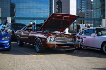 A classic muscle car with its hood open is parked amidst several other cars in a urban setting with tall buildings in the background. The car is reddish-brown and is displaying its engine as onlookers stand nearby, suggesting a car meet or show.