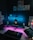 A modern computer setup includes a widescreen monitor and a laptop on a desk. The scene is illuminated with ambient lighting, featuring a blue glow behind the monitor and purple lighting under the desk. The monitor displays a nighttime landscape with a small illuminated building. The surroundings include various posters on the wall, speakers on either side of the monitor, a keyboard, mouse, and some books.
