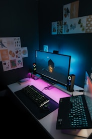 A modern computer setup featuring a large monitor with an image of a house in a mountainous landscape on the screen. The desk is illuminated by neon pink and blue lighting, which creates a futuristic atmosphere. A keyboard and mouse are positioned on the desk along with a laptop displaying an image. The walls are decorated with various art posters and designs.