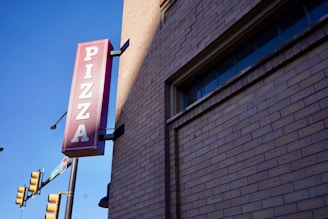 A red vertical pizza sign is attached to the corner of a building made of light brown bricks. The street corner is visible with traffic lights and a clear blue sky in the background.