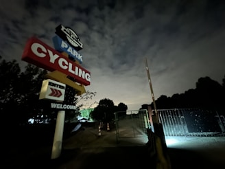 Evening shot of the bike park sign with warm lights glowing and trailheads fading into the dusk.