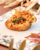 A golden-brown puff pastry filled with diced roasted vegetables and garnished with a single green sage leaf. The dish is presented on a decorative white plate with a black floral pattern and a hexagonal grid design. Autumn leaves and wheat stalks are softly blurred in the background, enhancing the cozy, seasonal atmosphere.