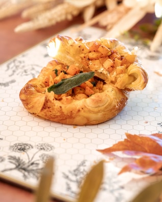 A golden-brown puff pastry filled with diced roasted vegetables and garnished with a single green sage leaf. The dish is presented on a decorative white plate with a black floral pattern and a hexagonal grid design. Autumn leaves and wheat stalks are softly blurred in the background, enhancing the cozy, seasonal atmosphere.