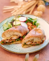 A plate with a sandwich cut into halves, held together by skewers, and filled with grilled chicken, cheese, and fresh greens. Accompanying the sandwich is a side salad containing mixed greens, radish slices, and cherry tomatoes. The setting includes a wooden table with soft-focus decorative elements in the background.