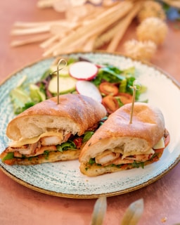 A plate with a sandwich cut into halves, held together by skewers, and filled with grilled chicken, cheese, and fresh greens. Accompanying the sandwich is a side salad containing mixed greens, radish slices, and cherry tomatoes. The setting includes a wooden table with soft-focus decorative elements in the background.