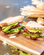 Two delicious-looking sandwiches placed on a wooden cutting board, filled with layers of crispy bacon, fresh green lettuce, and sliced avocado.