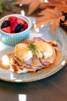 A dish featuring eggs Benedict on a waffle topped with a creamy hollandaise sauce and garnished with fresh dill. Ham is layered beneath the eggs. A side of fresh mixed berries, including strawberries, blackberries, and blueberries, is presented in a small blue ramekin. Autumn leaves are decoratively placed in the background, enhancing the fall theme.