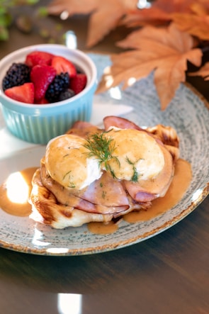A dish featuring eggs Benedict on a waffle topped with a creamy hollandaise sauce and garnished with fresh dill. Ham is layered beneath the eggs. A side of fresh mixed berries, including strawberries, blackberries, and blueberries, is presented in a small blue ramekin. Autumn leaves are decoratively placed in the background, enhancing the fall theme.