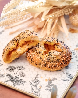 A close-up of a sesame and seed-covered bagel cut into halves, revealing a soft, cheesy filling inside. The bagel rests on a hexagonal-patterned surface with floral illustrations. In the background, there is a display of dried grasses and flowers, adding a rustic touch to the scene.