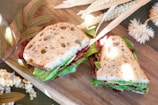 A sandwich on a wooden board, with layers of lettuce, avocado, and bacon between slices of rustic, holey bread. Surrounded by decorative dried leaves and flowers, creating a fresh and natural aesthetic.