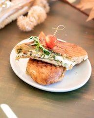 A grilled sandwich filled with creamy cheese and herbs sits on a white plate. The sandwich is toasted to a golden brown and garnished with a fresh basil leaf and a cherry tomato skewer.