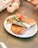 A grilled sandwich filled with creamy cheese and herbs sits on a white plate. The sandwich is toasted to a golden brown and garnished with a fresh basil leaf and a cherry tomato skewer.