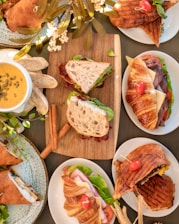An arrangement of various freshly prepared sandwiches on different types of bread, including croissants and paninis, displayed on white plates. Accompanied by a bowl of creamy soup garnished with pumpkin seeds and sliced baguette. Decorative elements include green leaves and flowers, adding a touch of elegance to the presentation.