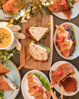 An arrangement of various freshly prepared sandwiches on different types of bread, including croissants and paninis, displayed on white plates. Accompanied by a bowl of creamy soup garnished with pumpkin seeds and sliced baguette. Decorative elements include green leaves and flowers, adding a touch of elegance to the presentation.