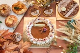 An assortment of pastries and baked goods arranged on a surface. The central item is a large round tart with a decorative rim and topped with small flowers and cinnamon sticks. Surrounding it are smaller tarts with pecans and powdered sugar, along with various types of bread. Decor includes brown and orange autumn leaves and white floral elements.