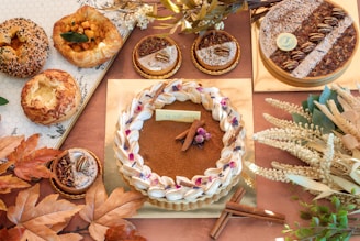 An assortment of pastries and baked goods arranged on a surface. The central item is a large round tart with a decorative rim and topped with small flowers and cinnamon sticks. Surrounding it are smaller tarts with pecans and powdered sugar, along with various types of bread. Decor includes brown and orange autumn leaves and white floral elements.
