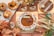 An assortment of pastries and baked goods arranged on a surface. The central item is a large round tart with a decorative rim and topped with small flowers and cinnamon sticks. Surrounding it are smaller tarts with pecans and powdered sugar, along with various types of bread. Decor includes brown and orange autumn leaves and white floral elements.