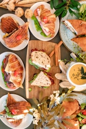 An assortment of sandwiches and soups are elegantly arranged on a table. The display includes croissant and ciabatta sandwiches filled with fresh greens, tomatoes, and various deli meats. Complementing the sandwiches are bowls of creamy soup garnished with seeds and herbs. The scene is decorated with dried flowers and greenery, creating a warm and inviting atmosphere.