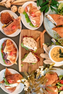 An assortment of sandwiches and soups are elegantly arranged on a table. The display includes croissant and ciabatta sandwiches filled with fresh greens, tomatoes, and various deli meats. Complementing the sandwiches are bowls of creamy soup garnished with seeds and herbs. The scene is decorated with dried flowers and greenery, creating a warm and inviting atmosphere.
