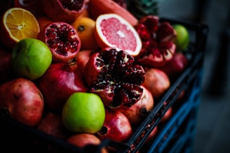 A collection of fresh fruits including pomegranates, a grapefruit, oranges, and green apples is displayed in a basket. The fruits are vibrant and juicy, with some pomegranates cut open to reveal their red seeds.