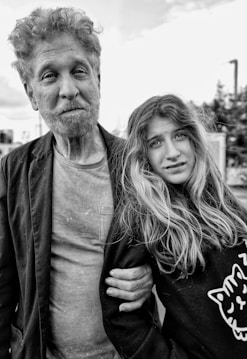 An older man and a young girl stand closely together. The man has curly hair and a beard, wearing a casual shirt and jacket. The girl, with long hair, looks at the camera. She wears a sweater with a cat illustration. Both have neutral to mildly serious expressions.