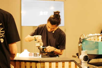 A barista is focused on preparing a cup of coffee, pouring milk with precision. The setting appears to be a modern coffee shop or a similar environment, with a coffee machine and various containers visible on a counter.