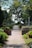 A serene cemetery scene with a brick pathway leading through well-maintained gardens. Large trees with abundant green foliage provide shade, while tombstones and graves are orderly arranged on either side. The sky peeks through the branches, casting soft light onto the path.
