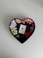 A heart-shaped box contains neatly arranged colorful ribbon rolls in a gradient from white and beige to various shades of pink and red. In the center of the box is a small white card featuring a printed red heart, accompanied by cursive text.