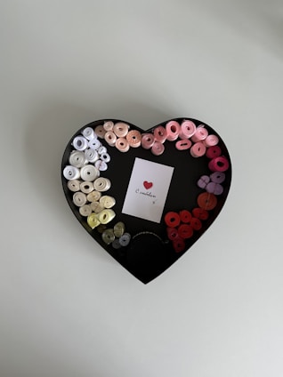 A heart-shaped box contains neatly arranged colorful ribbon rolls in a gradient from white and beige to various shades of pink and red. In the center of the box is a small white card featuring a printed red heart, accompanied by cursive text.
