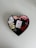 A heart-shaped box contains neatly arranged colorful ribbon rolls in a gradient from white and beige to various shades of pink and red. In the center of the box is a small white card featuring a printed red heart, accompanied by cursive text.