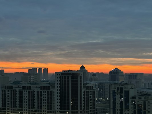 A panoramic cityscape at dusk with a vibrant orange and blue sky. Modern high-rise buildings dominate the foreground, silhouetted against the sunset. The skyline features a variety of architectural styles with several tall structures and a recognizable building with a dome.