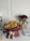 A bouquet of vibrant sunflowers and roses arranged with other colorful flowers, wrapped in burgundy paper with a cream ribbon. Nearby is a vase with red roses, a stack of papers, a pair of headphones, a glass, and a mug all set on a white table against a plain wall.
