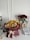 A bouquet of vibrant sunflowers and roses arranged with other colorful flowers, wrapped in burgundy paper with a cream ribbon. Nearby is a vase with red roses, a stack of papers, a pair of headphones, a glass, and a mug all set on a white table against a plain wall.