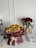 A bouquet of vibrant sunflowers and roses arranged with other colorful flowers, wrapped in burgundy paper with a cream ribbon. Nearby is a vase with red roses, a stack of papers, a pair of headphones, a glass, and a mug all set on a white table against a plain wall.
